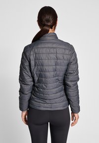 Vadderad grå jacka med ståkrage, quiltat mönster och långa ärmar, bärs över svarta leggings, sedd bakifrån.