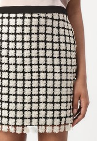 Jupe noire avec des fleurs blanches brodées ressemblant à des marguerites en motif quadrillé, portée par une personne avec une main visible sur le côté.