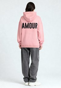 Felpa con cappuccio rosa con "AMOUR" stampato in nero sulla schiena, abbinata a pantaloni grigi larghi e sneakers bianchi. Tessuto morbido, vestibilità casual.