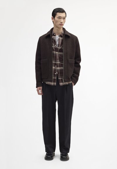 Braune Jacke aus Wollmischung mit Kragen, zwei Vordertaschen, über einem karierten Hemd getragen, kombiniert mit schwarzen, weit geschnittenen Hosen und Schuhen.