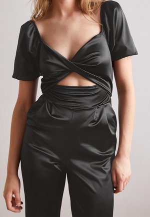 Kobieta ubrana w czarny satynowy top z bufkami na rękawach i skręconym przednim wycięciem, zestawiony z wysokim stanem czarnych satynowych spodni.