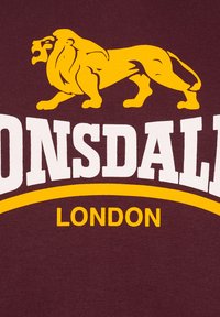 Felpa bordeaux con una grafica di un leone giallo. "Lonsdale" in lettere bianche in grassetto, con "London" in giallo sotto la grafica. Tessuto dalla texture liscia.