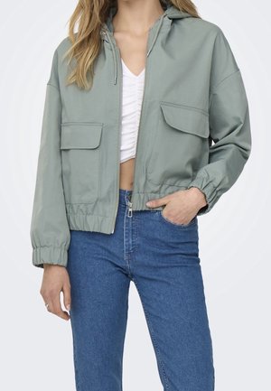 Femme portant une veste à capuche zippée vert clair avec de grandes poches devant, un crop top blanc froncé et un jean bleu taille haute.