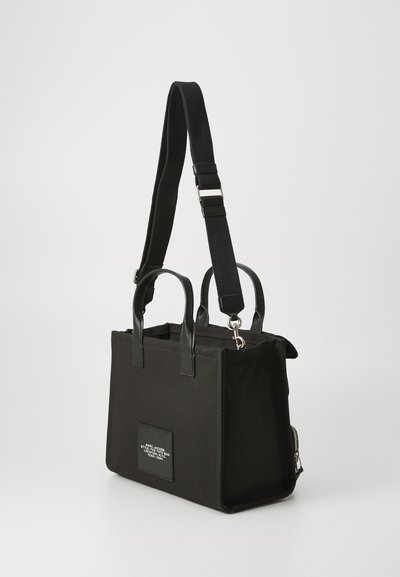 Black canvas handbag with dual top handles and an adjustable strap. Features a square patch with logo and a zippered side pocket.