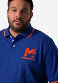 Modrá polokošile s oranžovým límcem a detaily, vyznačující se vyšitým logem "M+" a textem. Vyrobena z hladkého bavlněného materiálu.