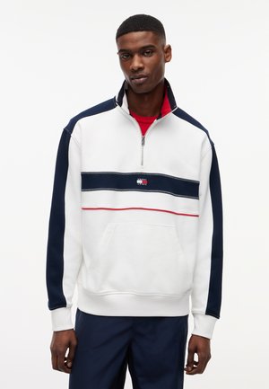 Młody mężczyzna ubrany w biało-granatową bluzę na suwak do połowy, z poziomymi paskami i logo, w połączeniu z granatowymi spodniami, stojący na białym tle.