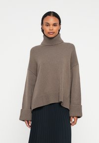 Femme portant un pull en col roulé taupe ample avec des manches retroussées et une jupe plissée noire, se tenant devant un fond blanc.