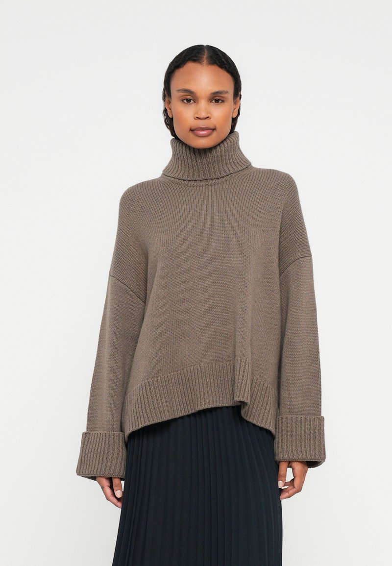 Femme portant un pull en col roulé taupe ample avec des manches retroussées et une jupe plissée noire, se tenant devant un fond blanc.