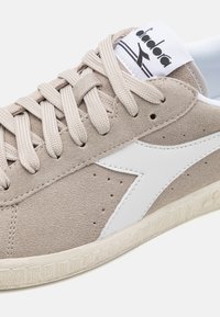 Beige suede sneaker with white leather accents, round toe, flat laces, and a textured rubber sole. Features a logo on the tongue.