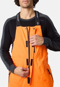 Heldere oranje waterdichte overall met verstelbare banden, rits aan de voorkant en zwarte accenten. Zacht zwart langepolssleeve top eronder.