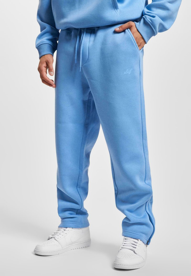 Hellblaue Sweatpants aus weichem Material, mit einem elastischen Bund, Seitentaschen und Reißverschlüssen an den Knöcheln. Kombiniert mit weißen Turnschuhen.