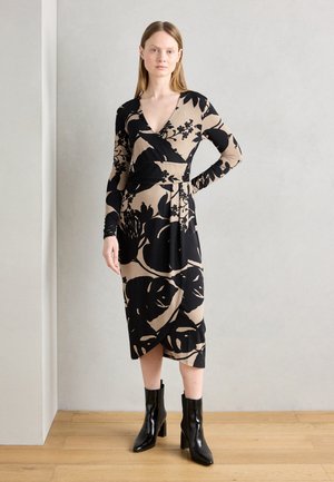 Maxi-jurk in beige met zwart bloemenpatroon, wikkelontwerp, lange mouwen, en enkel-lang. Gecombineerd met zwarte hoge laarzen.
