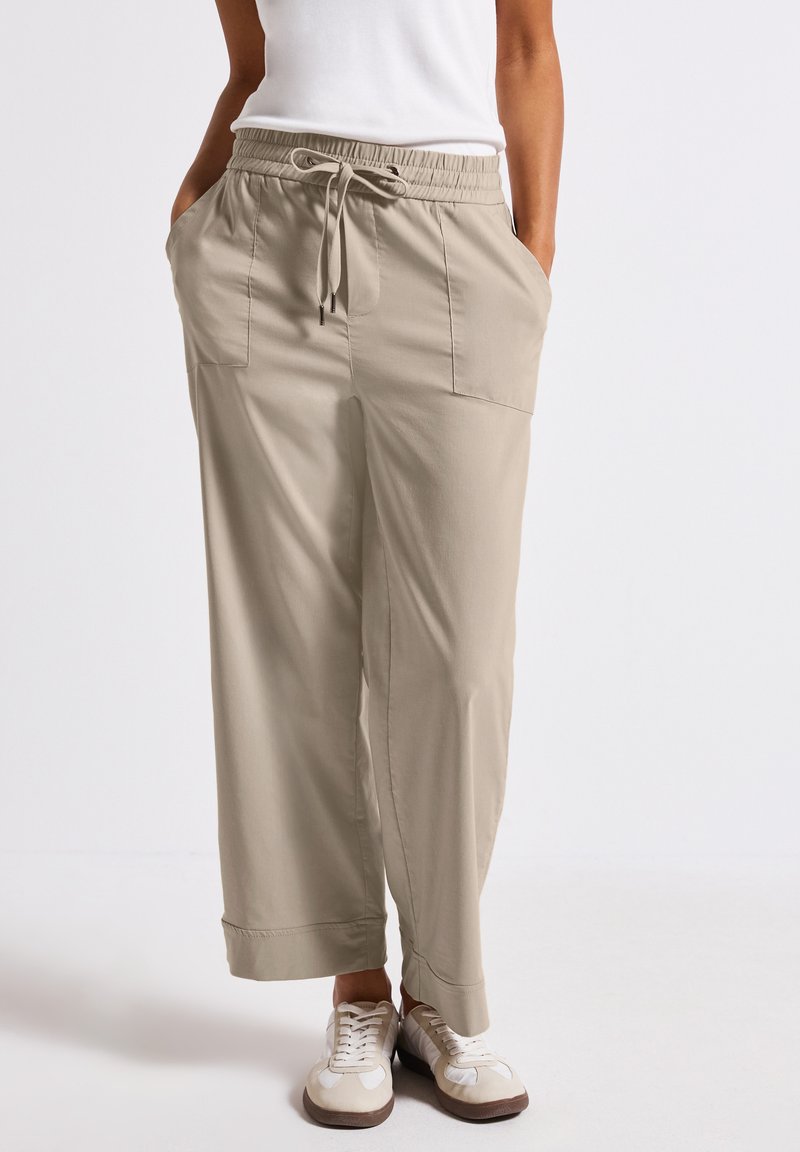 Lichtbeige, wijde broek met een elastische tailleband en trekkoord. Heeft twee voorzakken en een gladde, ademende stof.