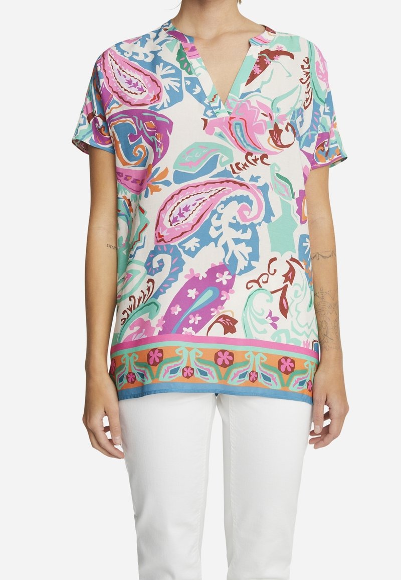 Femme portant un chemisier à manches courtes et col en V avec de grands motifs cachemire colorés et floraux, associé à un pantalon blanc.