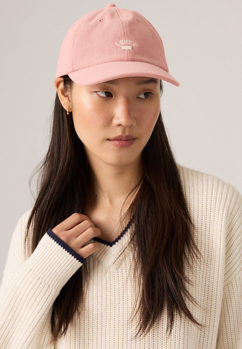 Pink corduroy cap with a curved brim and embroidered logo. Worn with a white knit sweater featuring dark blue accents.
