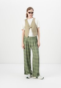 Kvinne som har på seg solbriller, beige vest over hvit t-skjorte, grønne rutete vide bukser og hvite joggesko, som står mot en hvit bakgrunn.