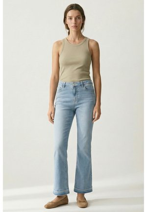 Frau steht frontal, trägt ein beiges Tanktop, hellblaue hochgeschnittene Jeans und beigefarbene flache Schuhe vor einem schlichten hellen Hintergrund.