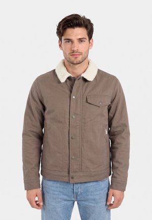 Hombre con chaqueta marrón con cuello de forro polar blanco y vaqueros azul claro, de pie frente a un fondo claro y liso.