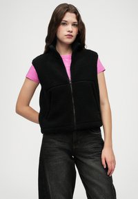 Svart fleecevest med høy krage og frontglidelås, brukt over en rosa skjorte. Har to frontlommer og en løs passform, kombinert med mørke jeans.