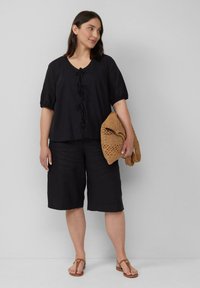 Schwarzes Kurzarm-Top mit V-Ausschnitt und dekorativen Vorderbindungen, kombiniert mit schwarzen Leinen-Culottes und einer geflochtenen braunen Tasche. Flache Sandalen vervollständigen den Look.