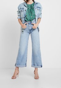Giacca di jeans azzurro chiaro sopra una blusa floreale verde, abbinata a jeans azzurro chiaro a gamba larga con orli arrotolati e tacchi nudi con cinturini.