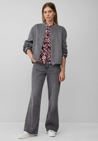 Vrouw staat met handen in haar zakken, gekleed in een grijze ritsjack, een gemarmerd bordeauxrood shirt, brede grijze jeans en witte sneakers tegen een effen achtergrond.