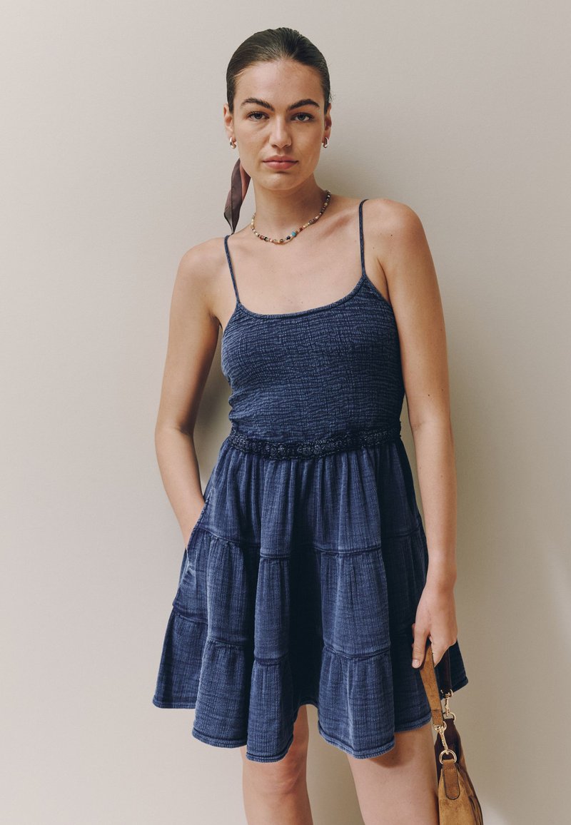 Jeune femme aux cheveux foncés attachés en arrière, portant une robe bleue texturée sans manches et tenant un petit sac à main marron sur un fond uni.