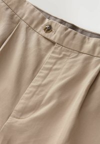 Beige Hosen mit glatter Textur, versehen mit einem mittigen Knopfverschluss, vorderen Falten und Seitentaschen. Dezente Nahtdetails sichtbar.
