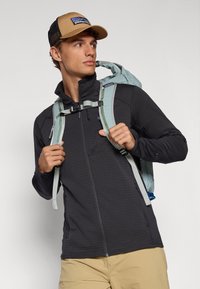 Black zip-up jacket with grid pattern, paired with a light blue backpack and beige cap; features visible zipper and adjustable straps.