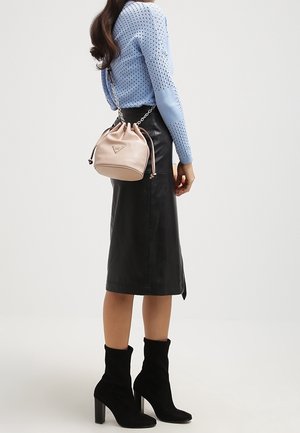 Femme portant un pull en maille bleu clair, une jupe midi en cuir noir, des bottines à talons noires, et tenant un sac seau en cuir beige avec une bandoulière chaîne.