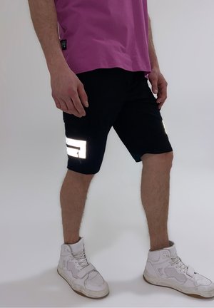 Uomo che indossa una maglietta fucsia, pantaloncini neri con strisce riflettenti vicino alle tasche e sneakers bianche alte, in piedi su uno sfondo chiaro.