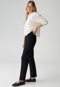 Femme portant une chemise blanche texturée boutonnée, un pantalon noir fuselé et des chaussures plates noires avec des brides à la cheville, debout devant un fond uni.