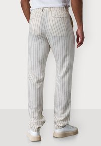Model trägt hellfarbene gestreifte Hosen und weiße Sneakers, von hinten gegen einen schlichten Hintergrund gezeigt.