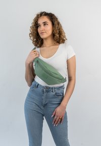 Borsa in tessuto verde con chiusura a zip, forma curva e patch rettangolare. Il modello indossa una camicia bianca e jeans azzurri.