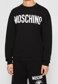 Černá mikina z měkké látky s velkým bílým logem "MOSCHINO" na přední straně. Klasický kulatý výstřih a uvolněný střih.