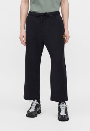 Persona che indossa pantaloni neri a gamba larga con bottoni automatici laterali, scarpe Nike nere e bianche, e una camicia verde chiaro infilata.