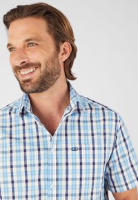 Camisa de botones de manga corta en un patrón de cuadros en azul, blanco y marino. Presenta un bolsillo en el pecho con logo y un diseño de cuello clásico.