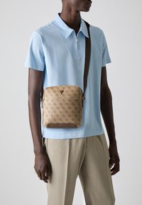 Sac bandoulière marron à motifs avec fermeture éclair, arborant un logo triangulaire. Associé à un polo bleu clair et à un pantalon beige.