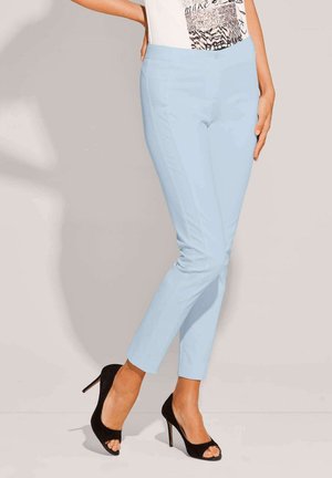 Woman wearing light blue slim pants, white graphic top, and black open-toe high heels, standing with one hand on hip.
