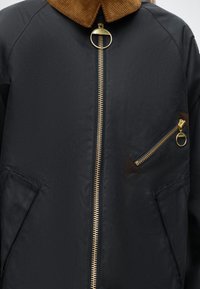 Veste noire avec grande fermeture éclair dorée à anneau, col en velours côtelé marron et poche zippée en biais sur la poitrine.