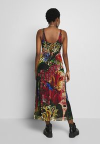 Kvinde med kort krøllet hår iført en ærmeløs blomster maxi kjole og sorte ankelstøvler, stående mod en ensfarvet grå baggrund.