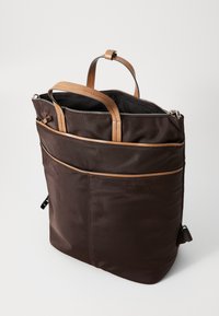Smeđa platnena torba s tamnocrvenim kožnim ručkama, zatvaračem na vrhu, prednjim džepom i elegantnim dizajnom; mekana tekstura, minimalistički stil.