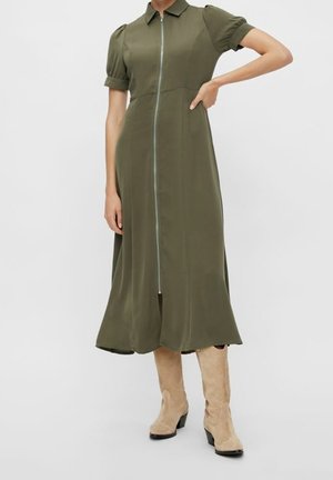 Vrouw draagt een olijfgroene midi-jurk met een rits aan de voorkant, pofmouwen en kraag, gecombineerd met beige suède laarzen tot halverwege de kuit, staand met één hand op de heup.