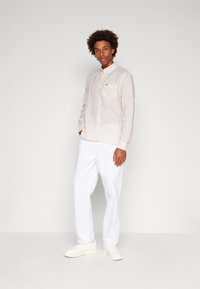 Camicia a maniche lunghe da uomo di colore beige chiaro con sottili righe verticali, dotata di un piccolo logo sul petto, abbinata a pantaloni bianchi.