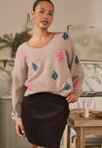 Lys grå strikket sweater med farverige træmønstre i pink og grøn; bæres med et sort mini-skjørt. Har en bred halsudskæring og lange ærmer.