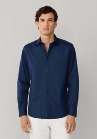 Granatowa koszula z długim rękawem, z guziczkami i kołnierzykiem, o gładkiej fakturze i dopasowanym kroju, zestawiona z białymi spodniami.