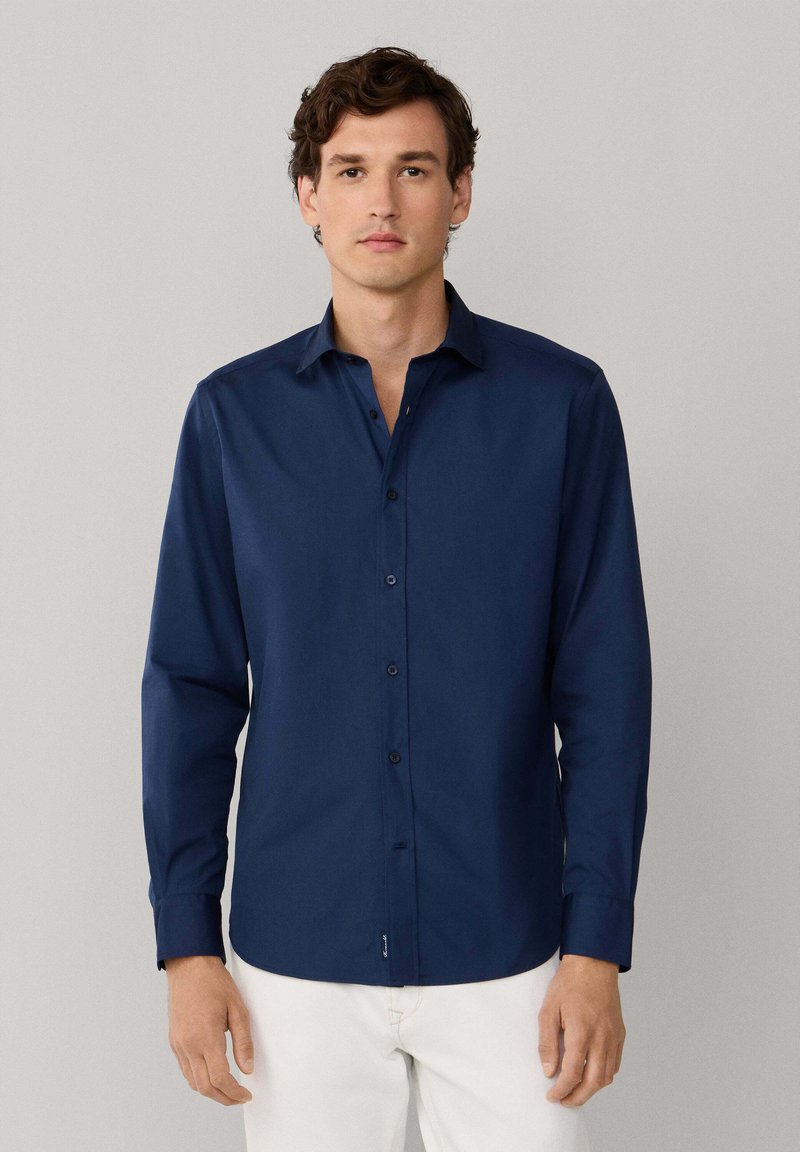 Granatowa koszula z długim rękawem, z guziczkami i kołnierzykiem, o gładkiej fakturze i dopasowanym kroju, zestawiona z białymi spodniami.