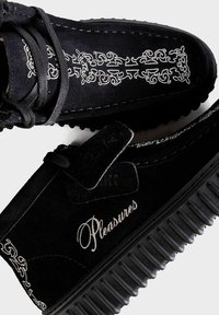 Schwarze Wildlederschuhe mit dicken, gerillten Sohlen, weißen bestickten Barockmustern und dem Wort "Pleasures" auf die Seite genäht.