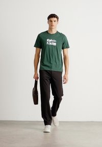 Young man walking wearing a green Calvin Klein T-shirt, black pants, white sneakers, and carrying a small black bag in right hand.