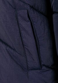 Materijal tamnoplave puffer jakne s teksturiranom završnom obradom, koja sadrži bočnu džep i kontrastne šavove.